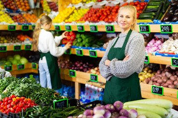 Confident successful female grocery store owner in apron standing near shelves of fruits and vegetables..