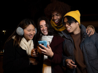 Group of diverse young people having a good time in winter clothes at night in a city while browsing the mobile phone