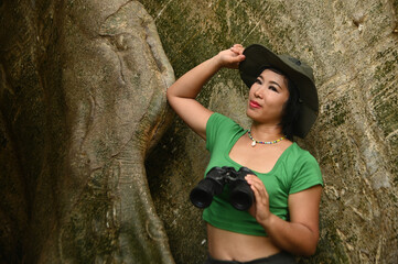 Pretty Asian woman Holding binoculars at the roots of a large tree and are delighted happily. This tree is very large and is a popular attraction for Thai tourists.