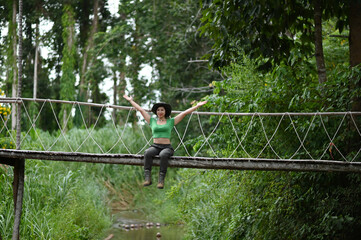 Fototapeta na wymiar Pretty Asian woman sitting and posing at the suspension bridge over the stream in the forest which is a tourist attraction happily.