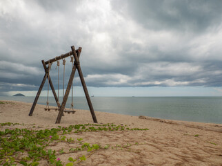 wooden swing tied with rope by the beach. beautiful tropical nature landscape. overlooking the bright green sea far and wide. the sky is cloudy with rain, and some are falling into the sea.