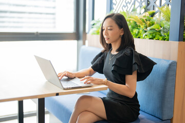 Short hair Asian female executive half body shot Company boss, wearing black suit, sitting cross-legged working with laptop about future business. The desk is in the office space.