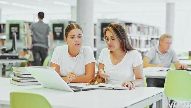 Positive female students in casual clothes using laptop and talking with each other in the book space library