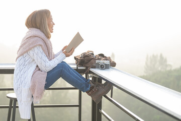 young woman reading book or bible near window and looking mountain view at countryside homestay in the morning sunrise with copy space. journey, trip and relaxing concept. coats blankets in winter. - Powered by Adobe