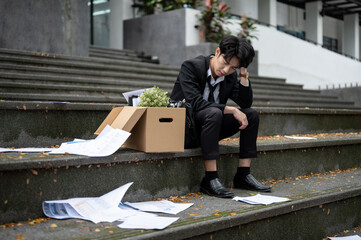 A sad Asian businessman sits on the stairs with a box of his personal stuff after losing his job