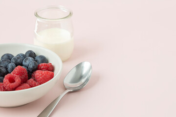 Closeup view of a white bowl with berries, glass of milk and a spoon on light pink background with copy space