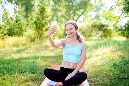 Young smiling sporty athletic fitness trainer instructor woman doing exercises do selfie shot on mobile cell phone sit on mat outdoor