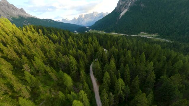 Dirt road surrounded by alpine forest in a valley in Cadore, Italy, with the Dolomites mountain range in the background at sunset. FPV drone shot. Base of Tre Cime di Lavaredo.