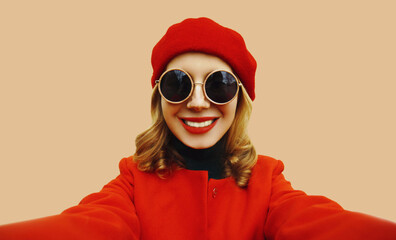 Autumn portrait of beautiful woman stretching hand for taking selfie with mobile phone wearing red french beret hat and round sunglasses on brown studio background