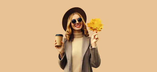 Autumn color style outfit, portrait of beautiful smiling young woman with yellow maple leaves and cup of coffee wearing round hat, coat on brown background