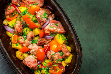 Turkey roast with potatoes, red paprika, onion, parsley, pepper and garlic in cast iron baking dish, rustic green table background, top view