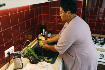 Side view of plus size black female of 60s washing vegetables with tap water at kitchen, holding...