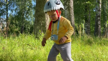 child in safety helmet with balance bike and toy camera on walk in summer park explores a mole hole