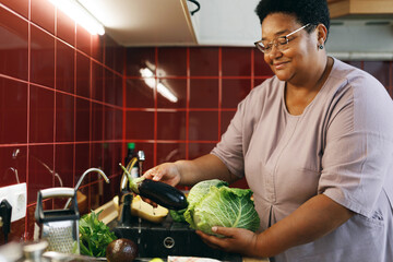 Happy plus size female of 60s with dark skin washing vegetables in sink with tap water, holding fresh aubergine and cabbage, cooking healthy dinner, enjoying process and harvest from her garden