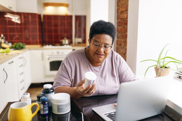 Plus size senior african american woman in glasses sitting at kitchen table full of food supplements in front of laptop, reading label and composition on vitamin bottle, working online as copywriter
