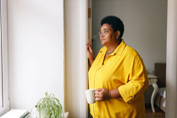 Side view of sad african american overweight female in yellow clothes and glasses standing next to window with cup of hot drink, grandma missing her grandchildren, waiting them to visit her - obrazy, fototapety, plakaty