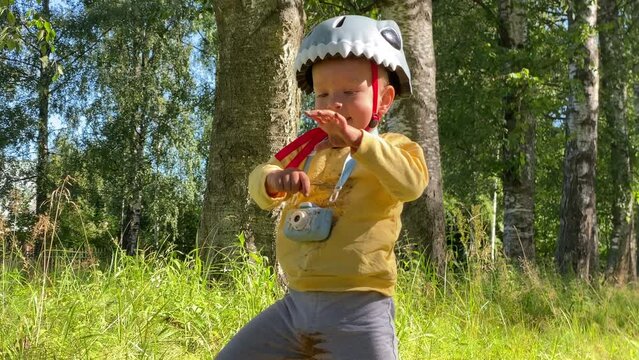 child in safety helmet with balance bike and toy camera on walk in summer park explores a mole hole