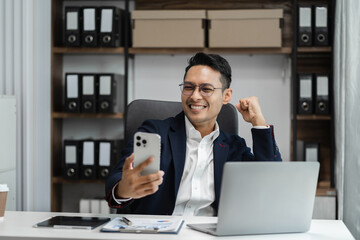 Obraz premium Excited man work distant on computer talk on cellphone celebrate online win. Happy young male use laptop feel euphoric overjoyed triumph with good news over smartphone call.