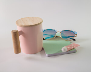 Taza de color rosa y gafas de color azul sobre una libreta color cafe y un brillo para labios