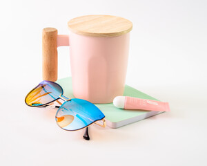 Taza de color rosa y gafas de color azul sobre una libreta color cafe y un brillo para labios