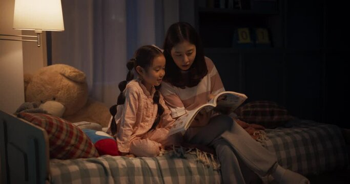 Korean Mother Reading a Fairytale to Her Lovely Small Daughter in Bed Before Going to Sleep. Young Beautiful Mom Caring for Little Girl, Reading Stories from a Book in the Evening at Home