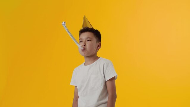 The asian kid boy in the paper cap folded her hands in a cross and she is very unhappy, disappointed and blows a party horn on plain yellow background