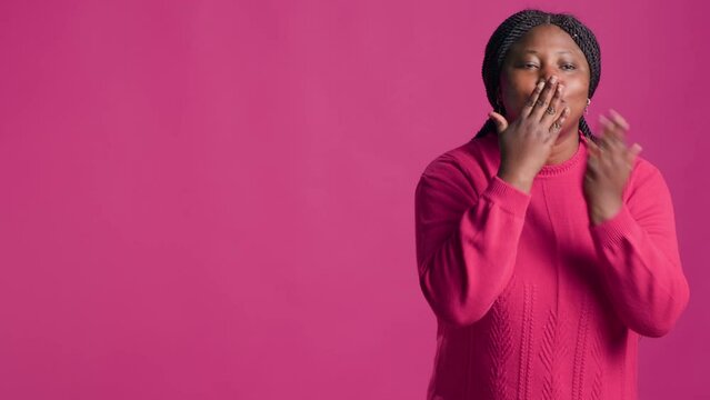 African american lady sending kiss towards camera while gracefully stands against isolated pink background. Stunning and joyful black woman conveys affection through cute hand gestures radiating love.