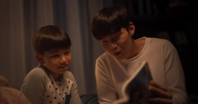 Korean Father Reading a Fairytale to His Lovely Cute Kids in Bed Before Going to Sleep. Patient Father Helping his Children to Learn how to Read Their Favorite Book. Parent and children Bonding Moment