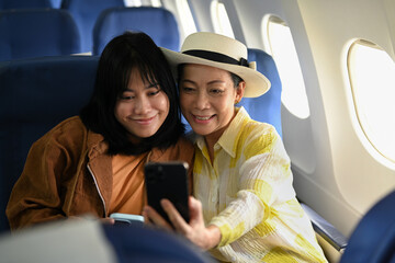 Happy 60s woman and daughter taking photo with smartphone in passenger airplane. Transportation,...