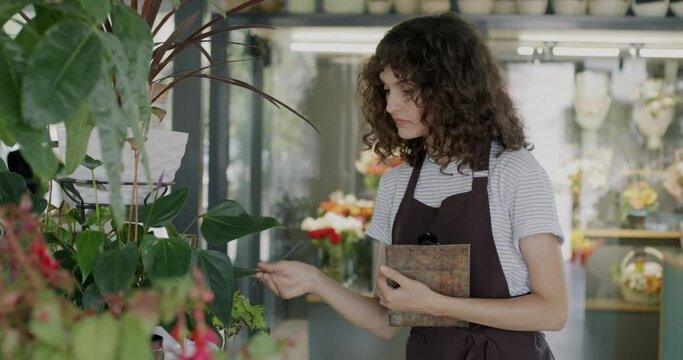 Young lady florist checking green plants and writing information in report working in modern flower shop. Commerce and floristry business concept.
