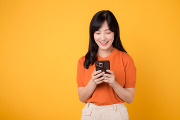 Smiling Asian woman in her 30s using smartphone, dressed in orange shirt, on vibrant yellow background. Mobile tech concept.