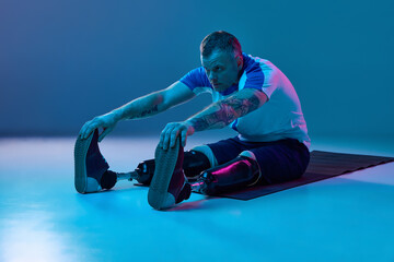 Confident man in sport uniform prosthetic legs doing stretching exercises on floor in neon light. Motivation poster.