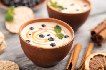 Yoghurt with bilberry and caramel in clay bowl on gray wooden, side view, selective focus.