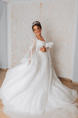 Fototapeta premium A happy bride is preparing for her luxurious wedding at home, holding her beautiful dress in her hands. Portrait of a woman with professional hair and makeup and a smile in a bathrobe