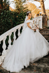 Fototapeta premium Portrait of the bride in the autumn park on stone steps. The bride in a wedding dress on a natural background with a bouquet of flowers in her hands. Wedding day.
