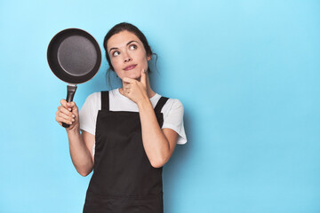 Woman with apron and pan on blue background looking sideways with doubtful and skeptical expression.