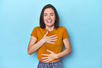 Young caucasian woman on blue backdrop laughs happily and has fun keeping hands on stomach.