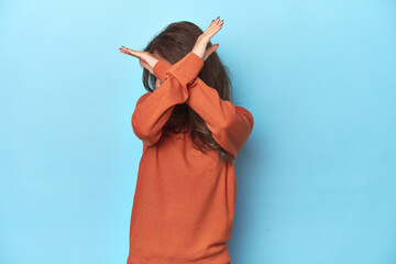 Teen girl in vibrant orange sweater on blue keeping two arms crossed, denial concept.