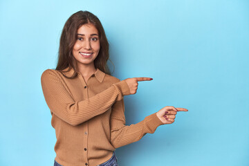 Teen girl in brown polo shirt on blue pointing with forefingers to a copy space, expressing excitement and desire.