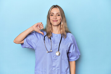 Young caucasian nurse in blue studio showing a dislike gesture, thumbs down. Disagreement concept.