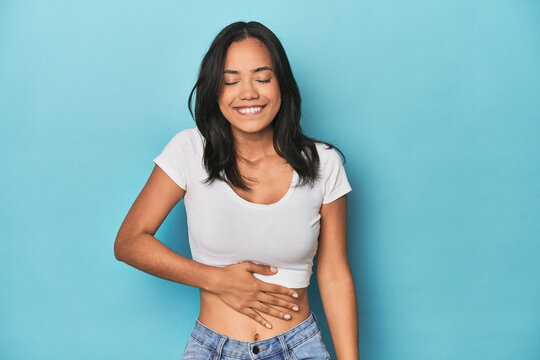 Filipina young woman on blue studio touches tummy, smiles gently, eating and satisfaction concept.