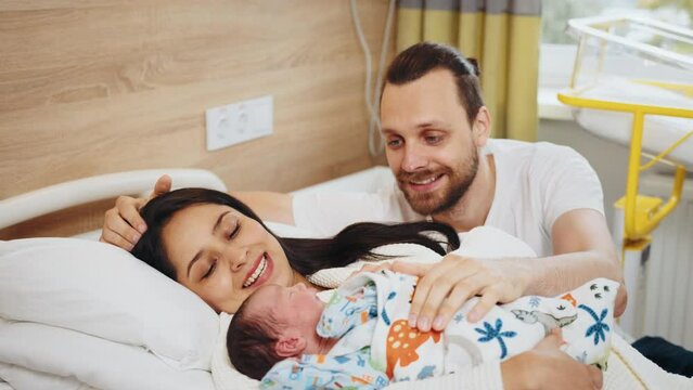 Side view of multiethnic couple looking at their new born baby at hospital ward. Father mother hugging on hospital bed look with love their son at post natal department