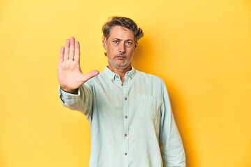 Middle-aged man posing on a yellow backdrop standing with outstretched hand showing stop sign,...
