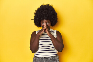 Obraz na płótnie Canvas African-American woman with afro, studio yellow background holding hands in pray near mouth, feels confident.