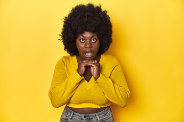 Fototapeta na wymiar African-American woman with afro, studio yellow background praying for luck, amazed and opening mouth looking to front.