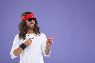 Stylish hippie man in sunglasses pointing at something on violet background, space for text
