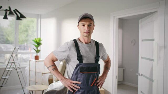Repairman taking hands to sides. Happy repair master standing at renovated apartment. Industry man wearing uniform and cap looking at camera. Satisfied craftsman smiling in flat after repair work.