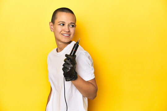 Tattooist woman in studio, shaved head, yellow background looks aside smiling, cheerful and pleasant.