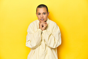 Young woman in jacket, yellow studio backdrop praying for luck, amazed and opening mouth looking to front.