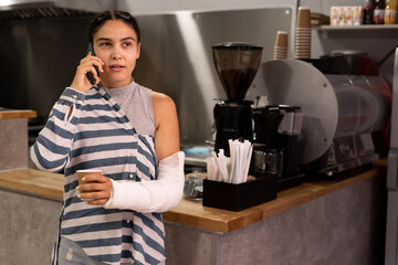 Young woman with a broken arm in a cafe drinking coffee talking on a mobile phone
